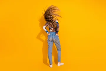 Young woman with flowing hair stands against a bright yellow background wearing jeans and a tie dye shirt © deagreez