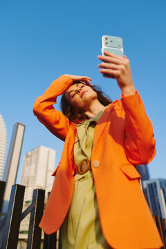 Sunscreen product promo, young woman in orange blazer squints in bright sunlight, shielding her face with hand while taking selfie against blue city sky on sunny day.