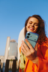 Beauty influencer, smiling young woman with long hair takes a selfie on her smartphone against a blue sky and modern city buildings, making direct eye contact with the camera.