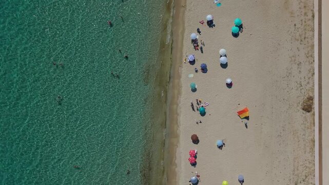 Drone footage shows a sandy beach with rows of umbrellas, swimmers and clear turquoise shallow sea along the shoreline.