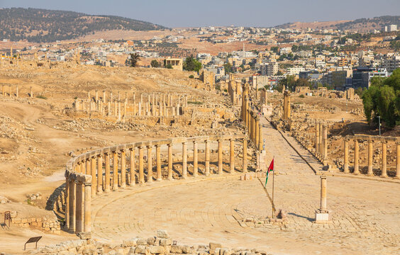 Aerial view of the ancient Roman ruins of Jerash, featuring the iconic Oval Plaza and the long Colonnaded Street, set against the modern city landscape in Jerash, Jerash Governorate, Jordan.