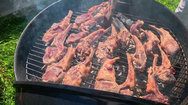 Portioned lamb chops (rack of lamb) are grilling on the grate of a round charcoal grill.