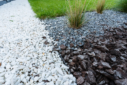 Split garden bed shows white pebbles, gray crushed stone, and dark bark mulch bordered by crisp edging, with ornamental grass clump and trimmed lawn in view.