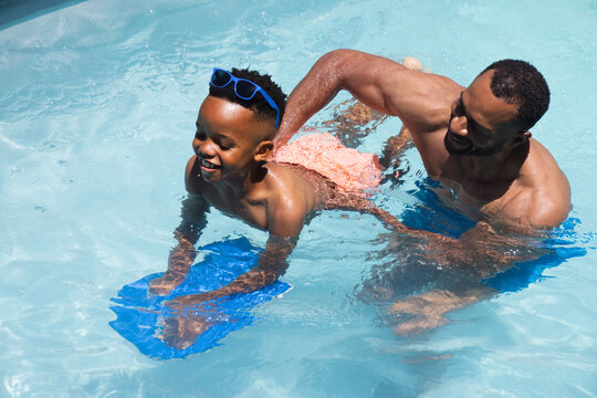 African American dad and schoolboy practicing swimming in pool, supporting blue kickboard, goggles