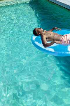 African American man reclining on blue ring, floating in turquoise pool in pink trunks, copy space