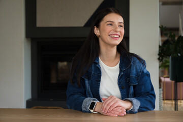 Naklejka premium Woman sitting at wooden table smiling and looking right wearing denim jacket and smartwatch