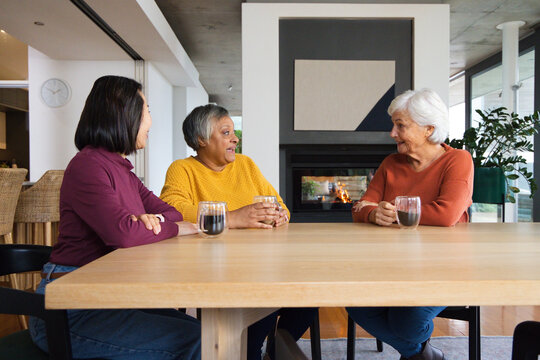 Diverse female friends sitting and chatting at dining table holding glass mugs by fireplace