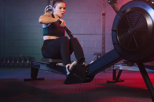 Woman rowing on rowing machine at gym, wearing dark sports bra and leggings, gripping handle