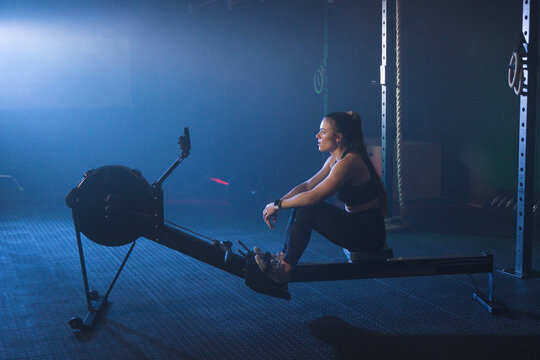 Female athlete sitting on rowing machine at gym, using phone mount and rope, copy space