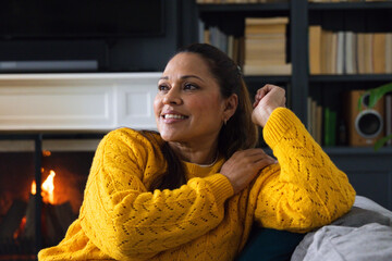 Fototapeta premium Middle-aged African woman resting on couch by lit fireplace, wearing mustard sweater