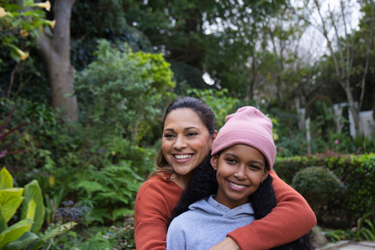 African American mother hugging teenage daughter, smiling in backyard garden with pink beanie