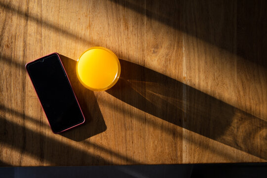 Glass of orange juice is sitting on wood table by window, smartphone lying, casting slatted shadows