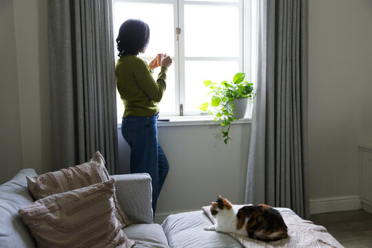 African American woman standing at bright window holding ceramic mug with plant and cat nearby