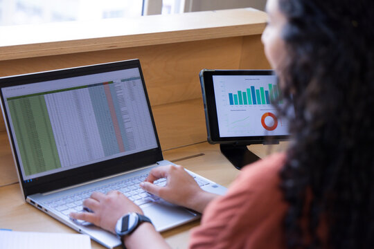 African American woman typing on laptop at desk, viewing tablet charts, rust top, watch, copy space