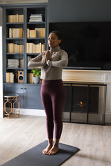 Fototapeta premium African adult woman in athletic clothes practicing yoga on black mat in living room by fireplace