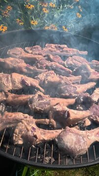 Portioned lamb chops (rack of lamb) are grilling on the grate of a round charcoal grill.