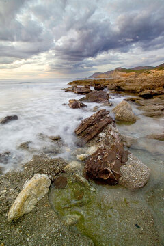a rugged, weathered rocky shoreline with small tidal pools reflecting a partly cloudy sky and gentle waves rolling into the sea under a soft daylight. Argentiera, Sardinia, Italy