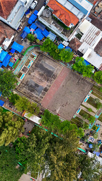 Aerial view of a basketball court surrounded by lush trees, vibrant market stalls with blue tarps, and residential rooftops in Ciwidey, Bandung, West Java, Indonesia.