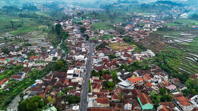 Aerial view of Ciwidey town featuring a central road, dense residential housing with red rooftops, and surrounding terraced rice paddies in Bandung, West Java, Indonesia.