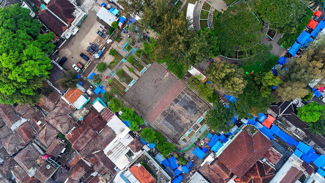 Aerial view of Ciwidey town square featuring a basketball court, surrounded by dense residential rooftops, lush trees, and vibrant blue-tarp market stalls in Bandung, West Java, Indonesia.