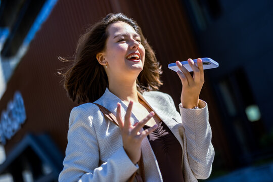 Expressive businesswoman holding smartphone and talking on speakerphone outdoors