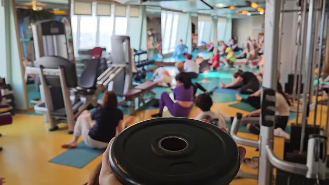 SOCHI, CRUISE - APR 9, 2026: Dumbbell held in foreground. Group exercises near large windows. Ocean view gym equipment. People work out on mats. Fitness room . Adults engage in workout routines