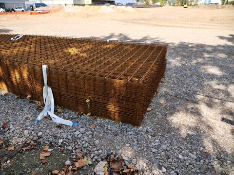 Stack of Rusty Steel Rebar Mesh Grid Panels Secured with White Straps at Outdoor Construction Site on Gravel Ground