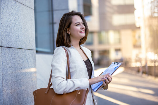 Smiling young businesswoman holding clipboard and handbag outdoors near office