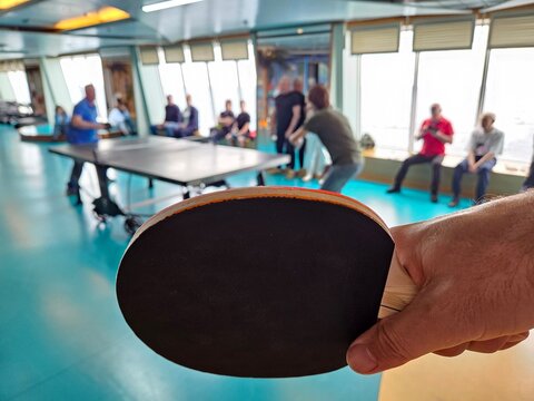Hand holds ping pong paddle in foreground. Players engage in table tennis match behind. Group gathered around multiple tables. Bright room with large cruise windows. Floor has blue and light sections