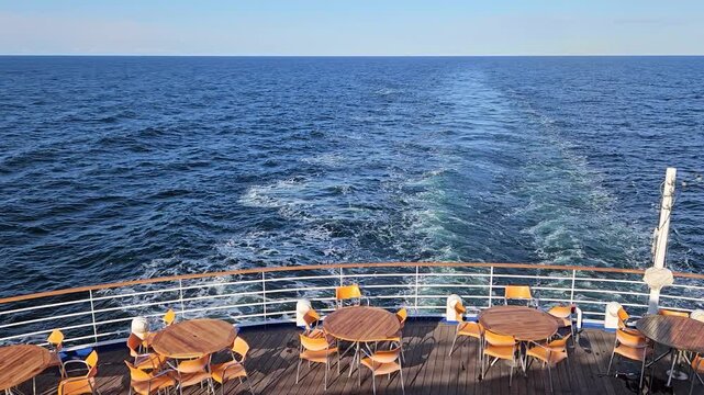 Cruise ship deck with wooden tables and chairs. Blue ocean stretches beneath clear sky. Waves trail behind vessel wake. Sunlight glints off calm sea surface cruise