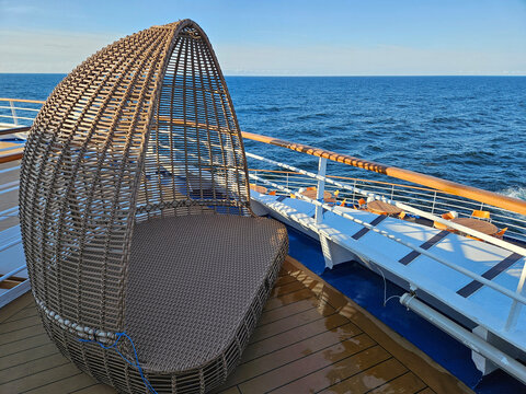 Woven egg-shaped chair on cruise ship deck. Ocean stretches under clear blue sky. Wooden deck with metal railings beside chair. Sunlight highlights the chair woven texture