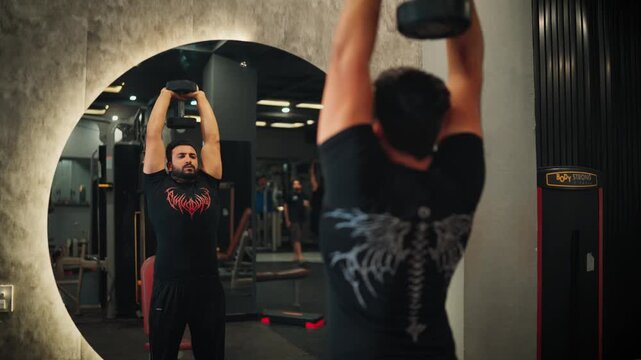 Muscular male bodybuilder executing overhead dumbbell triceps extension exercise targeting upper arm strength and triceps muscle hypertrophy in a modern fitness center.