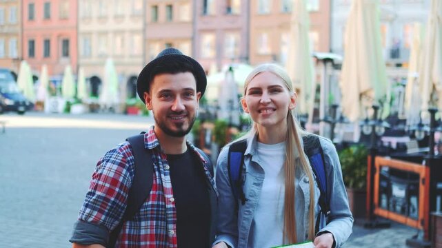 Two young tourists, a man and woman, smiling and looking at a map while standing in a busy European market square during daytime.