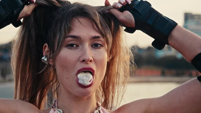 Close up shows young skater girl blowing bubblegum fixing hair. Beautiful sporty female wears protective wrist guards looking directly into camera during warm sunset.