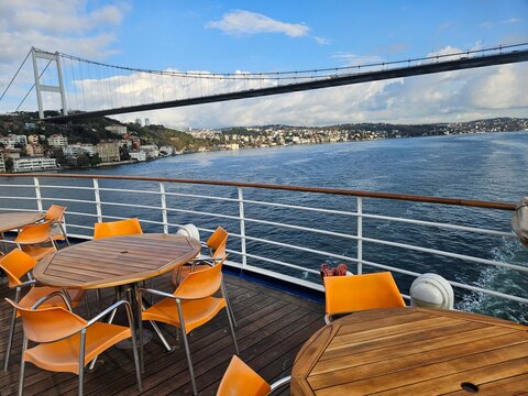 Boat deck offers ocean views with vibrant orange chairs and round tables. Bridge stretches across water beneath blue sky. Cityscape looms in distance, framed by the vessel railing in Istanbul, Turkey