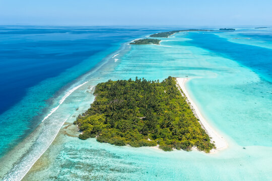 Aerial view of a tiny island with lush vegetation and white sand beach, surrounded by a turquoise lagoon and deep blue ocean in Vaanee, Dhaalu Atoll, Maldives.