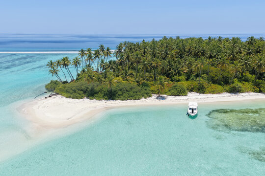 Aerial view of a tropical island with lush palm trees, white sand beach, and a small boat anchored in the clear turquoise lagoon in Vaanee, Dhaalu Atoll, Maldives.