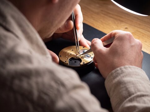 Watchmaker using precision tweezers to repair a mechanical watch movement at his workshop table under bright lamp light