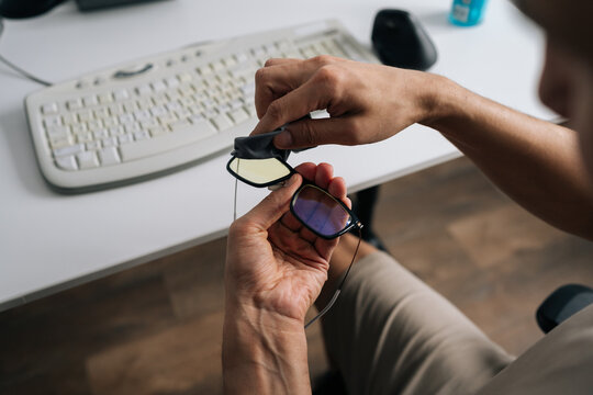 Overhead view of male hands carefully wiping blue light blocking glasses with microfiber cloth. Man taking break from work at office desk to clean eyewear for better vision and eye care.