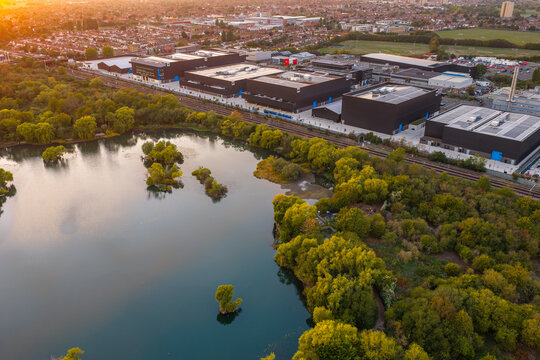 Aerial view of NTT LON1B Data Centre complex situated next to a tranquil lake with lush trees and railway tracks during golden hour in Dagenham, England, United Kingdom.