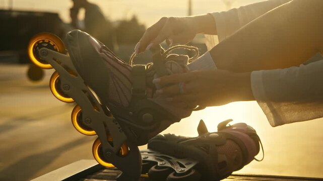 Close up of female hands fastening inline skates straps at sunset. Warm golden hour sunlight illuminates process while people play basketball in blurred background at urban extreme park.