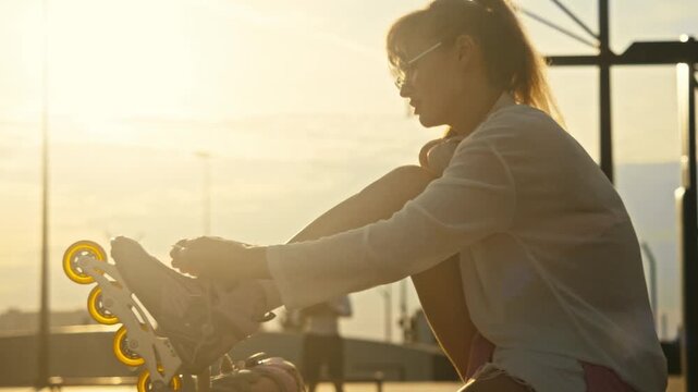 Medium shot of young woman in glasses putting on roller skates. Warm evening sunlight surrounds female skater preparing for active outdoor workout at urban sports park.