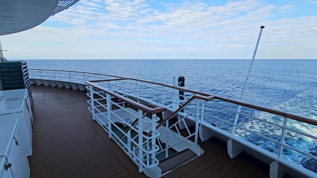 Cruise ship sails across open ocean. Deck view shows railing and wake. Sky is blue with scattered clouds. View from upper deck, calm sea. No animals or plants visible