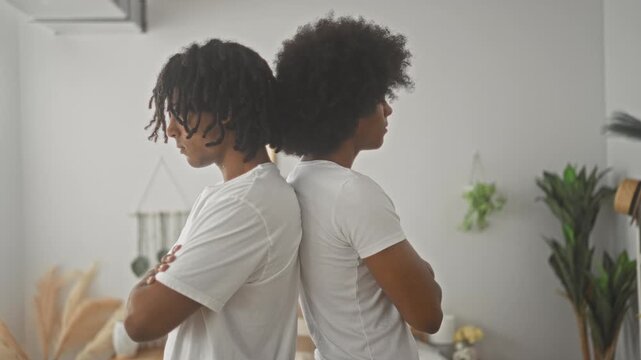Two african american men stand back to back with arms crossed, wearing white t shirts in a bedroom studio; friendship distance tension.