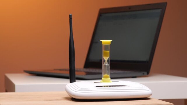 Hourglass with sand falling placed on a wireless internet router, symbolizing slow network connection and waiting for data to load on a blurred laptop screen in the background