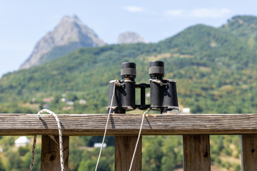 Binoculars rest on a weathered wooden railing facing a lush mountain view, evoking travel, exploration and outdoor observation. Perfect for hiking, birdwatching, scenic landscape and adventure themes © evannovostro