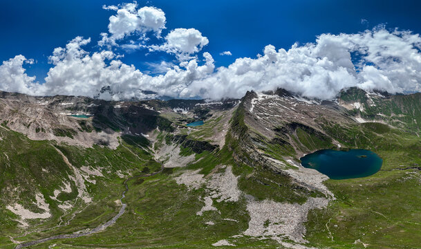 Aerial view of Maheen Lake surrounded by rugged mountain peaks, lush green alpine meadows, and rocky terrain under a blue sky with clouds in Batakundi, Khyber Pakhtunkhwa, Pakistan.