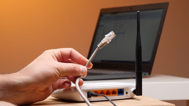 Hand holding an ethernet cable, preparing to connect it to a white wireless router with a laptop computer in the background, illustrating home internet connectivity and network setup