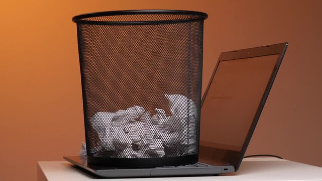 Mesh wastebasket overflowing with crumpled white paper sitting on an open laptop keyboard, symbolizing discarded ideas, failed projects, or digital cleanup