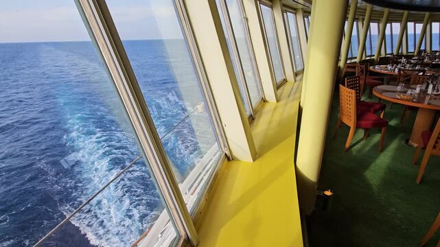 Bright yellow deck near ocean window. Large cruise window frames blue sea and wake. Green carpet and wooden tables inside. Sunlight glows through glass panes.